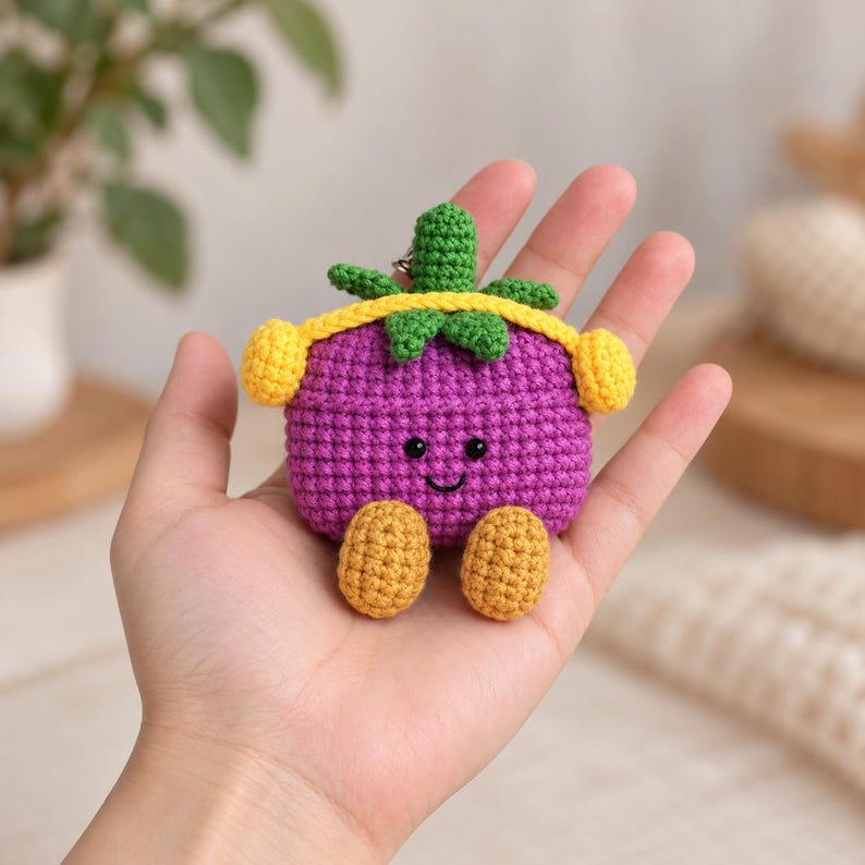 May include: A purple crochet case featuring a smiling face with black eyes and a stitched mouth. The case has a green leafy top, a yellow strap across the middle, two yellow side accents, and two small yellow feet at the bottom.