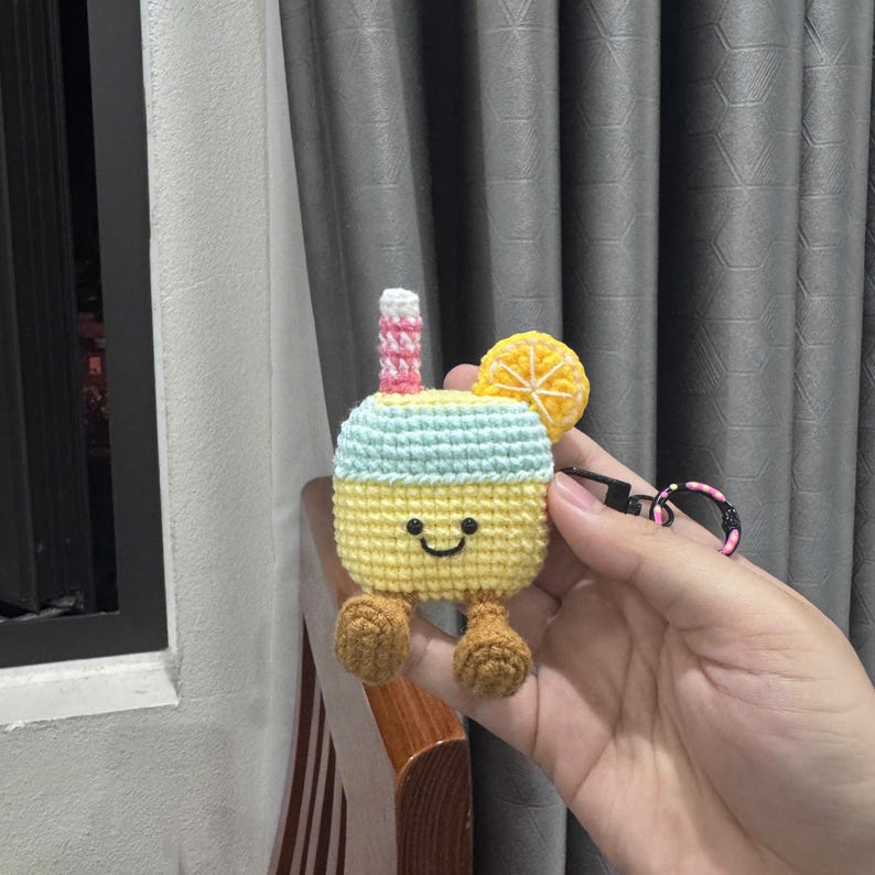 May include: A crochet case for wireless earbuds shaped like a drink with a yellow base, light blue top, a pink and white striped straw, and a yellow citrus slice. The case features a smiling face, two brown feet, and a black metal keychain attachment.