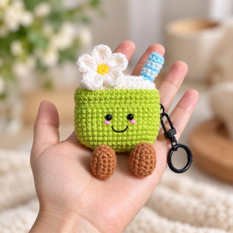 May include: A lime green crochet case for wireless earbuds featuring a smiling face, two brown feet, a white flower with a yellow center on top, and a blue and white striped straw. A black metal keychain ring is attached to the side.