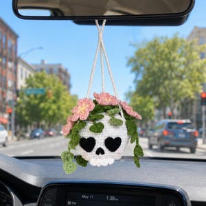 May include: A white crocheted skull hanging ornament with heart-shaped black eye sockets and a small nose opening. The skull is topped with green crocheted vines and several pink flowers, suspended by two white yarn strings from a car mirror.