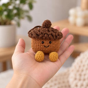 May include: A small, crocheted acorn-shaped case with a dark brown textured cap and a light brown body. It features two small black bead eyes, a stitched black smile, and two light yellow round feet attached to the bottom.