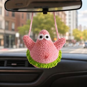 May include: A pink crocheted plush figure resembling a starfish with large white eyes and black pupils, wearing a textured green skirt. The figure has two pink yarn loops extending from the top, designed to hang from a vehicle rearview mirror.