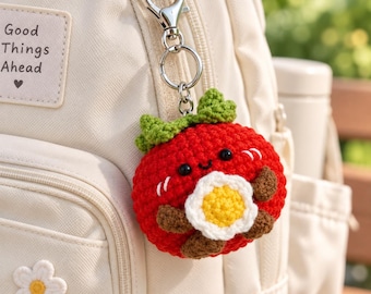 May include: A crochet keychain featuring a round red character with green leaves on top, black eyes, and a small smile, holding a white and yellow flower. The keychain is attached to a cream-colored backpack with a patch that reads Good Things Ahead.
