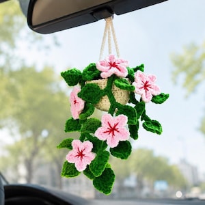 May include: A crocheted hanging plant ornament featuring a small cream-colored pot, vibrant green leaves, and five-petaled light pink flowers with dark red centers. The plant is suspended by a cream-colored yarn cord from a vehicle rearview mirror.