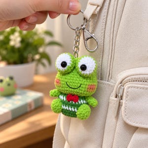 May include: A crochet frog keychain featuring a green body, large white eyes with black pupils, pink cheeks, and a small red bow tie. The frog wears a green and white striped shirt and is attached to a silver metal keyring and lobster clasp.