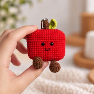 May include: A red crochet case for wireless earbuds shaped like an apple with a smiling face, black bead eyes, a brown stem, a green leaf, and two small brown dangling feet attached to the bottom, featuring a silver metal keyring loop.
