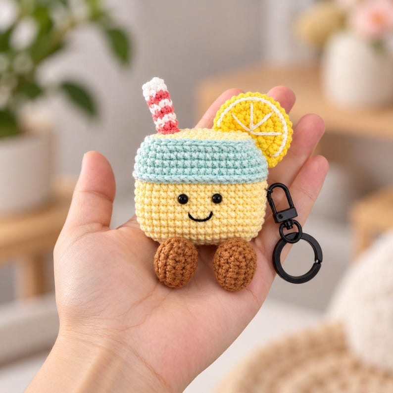 May include: A crochet case for wireless earbuds shaped like a drink with a light blue top, yellow base, and brown feet. It features a red and white striped straw, a yellow lemon slice garnish, and a black metal keychain ring attached to the side.