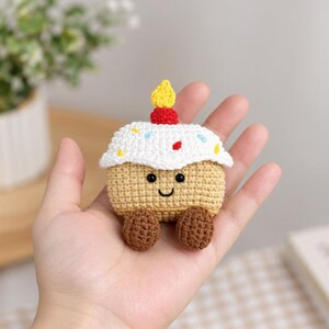 May include: A small, tan crocheted cupcake toy with a white frosting top, colorful sprinkles, a red cherry, and a yellow candle. The cupcake features a smiling face with black bead eyes and two small brown feet at the base.