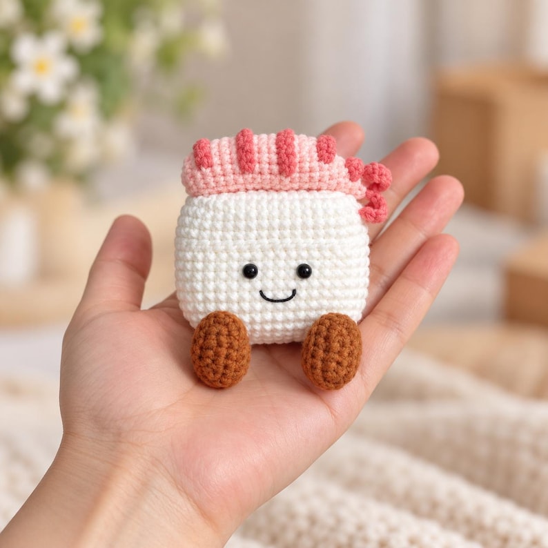 May include: A small, handmade crochet plushie shaped like a piece of shrimp sushi. The item features a white rice base, a pink shrimp topping, and two brown feet. It has a stitched black smile and two small black bead eyes on the front.