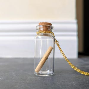 May include: A clear glass bottle necklace with a cork stopper and a gold-colored chain. Inside the bottle is a rolled-up paper scroll. The bottle is approximately 2 inches tall.