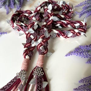 May include: Braided burgundy and white hair accessory with silver leaf accents and decorative charms. The accessory features multiple strands of braided fabric, with silver leaf embellishments and decorative charms at the ends. The overall design is intricate and elegant.