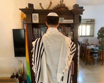 Puede incluir: Una persona que lleva un talit blanco con rayas negras y una kipá blanca, de pie frente a una estantería de madera. Una menorá y otros objetos religiosos están encima de la estantería. La persona está de espaldas a la cámara.