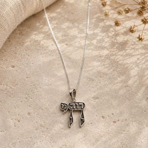 May include: A silver necklace with a pendant in the shape of the Hebrew letter "Chai". The pendant has an openwork design with Hebrew letters. The necklace is displayed on a neutral-toned surface with a soft, textured cloth in the background.
