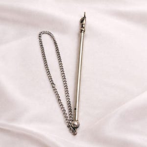 Może przedstawiać: Srebrny długopis z dołączonym łańcuszkiem. Pióro ma szczegółową końcówkę w kształcie dłoni skierowaną w górę. Łańcuszek ma delikatny, połączony wzór. Pióro jest prawdopodobnie przedmiotem dekoracyjnym lub kolekcjonerskim.