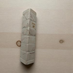 Może przedstawiać: Prostokątna, jasna obudowa mezuzy o marmurowym wyglądzie. Obudowa jest podzielona na prostokątne bloki i posiada złotą hebrajską literę. Tło to jasne usłojenie drewna.