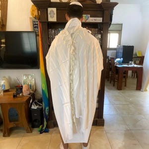 Op de afbeelding: Een witte tallit, een Joodse gebedssjaal, is over de schouders van een persoon gedrapeerd. De tallit heeft een decoratief, gestructureerd ontwerp op de rug. De persoon draagt een witte keppeltje. De achtergrond omvat een houten boekenkast en een televisie.