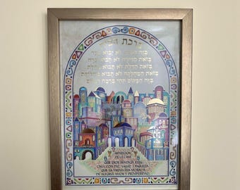 Puede incluir: Obra de arte enmarcada con una colorida ilustración de un pueblo con texto en hebreo y español. La obra de arte está en un marco de color dorado. El texto incluye una bendición para el hogar.