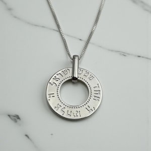 May include: A silver pendant necklace. The circular pendant has engraved Hebrew text and is suspended from a fine chain. The necklace is shown against a white marble background.
