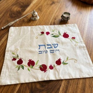 Könnte beinhalten: Ein quadratischer weißer Stoffbezug mit blauem hebräischem Text, der Shabbat Yom Tov lautet, umgeben von einer gestickten Girlande aus roten Granatäpfeln und grünen Blättern. Das Design ist mittig auf dem Stoff symmetrisch angeordnet.