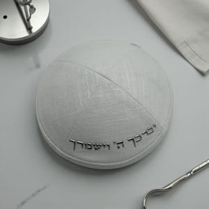 Può includere: Una kippah bianca con testo ebraico argentato. La kippah è rotonda e realizzata in un tessuto strutturato. Un oggetto in metallo argentato è sullo sfondo. La kippah è un tradizionale copricapo ebraico.