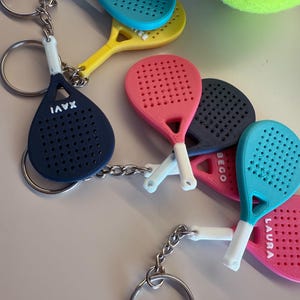 May include: Keychains featuring miniature padel rackets in various colors: blue, yellow, pink, and navy. Each racket has a white handle and a silver keyring. Some rackets have names printed on them, and a tennis ball is visible in the background.
