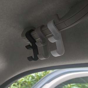 Puede incluir: Tres ganchos interiores para coche en negro, gris y blanco están sujetos a un panel de techo gris. Los ganchos están diseñados para colgar objetos dentro de un vehículo. Los ganchos son de plástico.