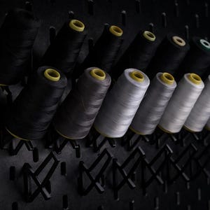 May include: A collection of spools of thread in various shades of black, gray, and white, with yellow cores. The spools are mounted on a black pegboard, creating a visually organized display for sewing or crafting projects.