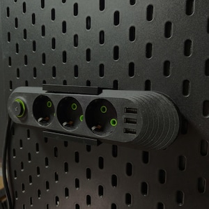 May include: A black power strip with three electrical sockets and two USB ports is mounted on a black pegboard. The sockets have green accents, and a green power button is visible.