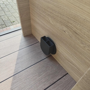May include: A black, plastic, dual-compartment holder fixed to a light wood-grain cabinet. The holder is mounted near the floor, with a circular base. The cabinet is made of light-coloured wood with visible grain.