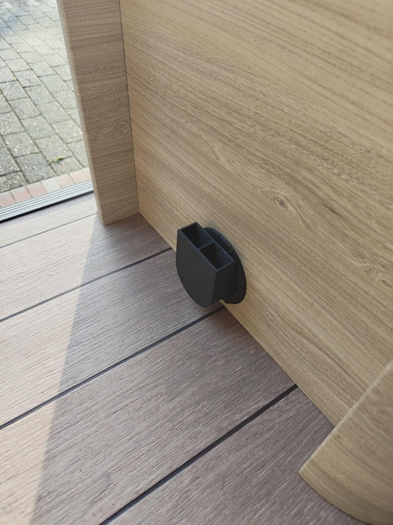 May include: A black, plastic, dual-compartment holder fixed to a light wood-grain cabinet. The holder is mounted near the floor, with a circular base. The cabinet is made of light-coloured wood with visible grain.