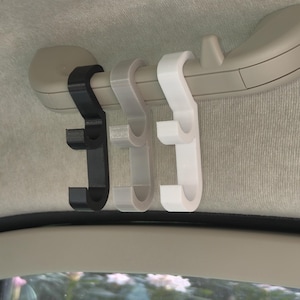 Op de afbeelding: Drie hoofdsteunhaken voor in de auto in zwart, grijs en wit. De haken zijn bevestigd aan een lichtgrijze stang, ontworpen om items op te hangen. De haken zijn gemaakt van plastic en zijn ontworpen om in een auto te passen.