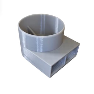 May include: A light grey 3D-printed object. It has a cylindrical top and a rectangular base with two openings. The cylindrical part has a divider inside. The object is made of plastic and shows layer lines from the printing process.