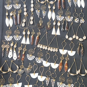 May include: A large collection of assorted dangle earrings featuring gold-tone wire, white cowrie shells, and black and white patterned geometric shapes. The earrings include various designs such as triangles, spirals, and teardrop forms.