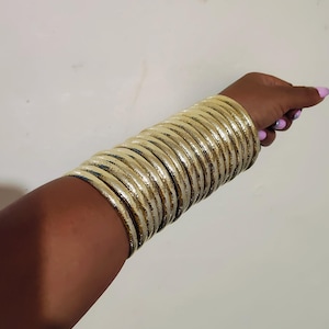 Op de afbeelding: Een verzameling goudkleurige armbanden die op een arm zijn gestapeld. De armbanden hebben een textuur en worden samen gedragen, wat een gedurfde uitstraling creëert. De arm staat tegen een effen witte achtergrond.