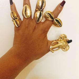 May include: A collection of five gold-toned rings worn on fingers. Four rings feature cowrie shell designs with dark central slits, while the fifth ring on the thumb displays a detailed elephant head motif with a trunk extending downward.