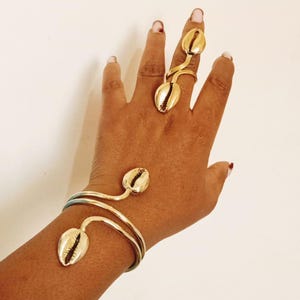 May include: A gold-coloured jewellery set comprising a ring and a bracelet, both decorated with seashell-shaped details. The ring has two shells, and the bracelet has one. The jewellery is shown on a hand against a white backdrop.