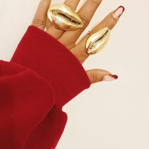 May include: Two gold-colored shell-shaped rings are worn on a hand with red nail polish. The rings are large and detailed, with a textured surface. The sleeve of a red sweater is visible in the image.