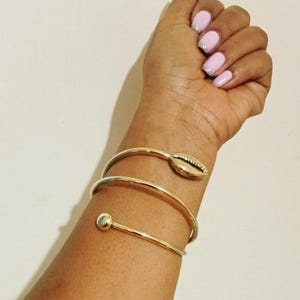 May include: Gold-coloured metal bracelet with a spiral design. The bracelet features a seashell accent and a small spherical detail. The arm is adorned with a gold bracelet and the nails are painted with a light pink colour.