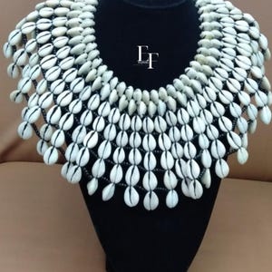 May include: A striking necklace made from numerous white and cream seashells, threaded onto black string. The necklace is displayed on a black mannequin bust. The shells are arranged in a layered, cascading design against a neutral background.