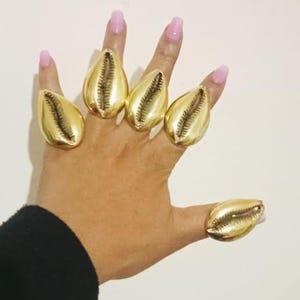 May include: Gold-colored cowrie shell rings. The rings are worn on the fingers, with a black sleeve visible in the corner. The background is a plain white surface. The nails are painted pink.