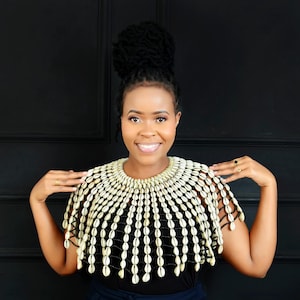 Puede incluir: Collar de conchas con cuentas sobre un fondo negro. El collar está hecho de conchas de color crema y hilo negro. Las conchas están dispuestas en filas, creando un efecto de capas. Una pieza de joyería llamativa.