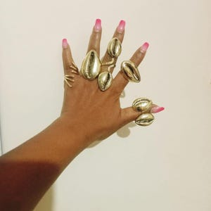 May include: A hand displaying several gold-colored rings. The rings feature a seashell design, with varying sizes and styles. The nails are painted with a pink ombre effect. The rings are the focal point, showcasing a unique jewelry collection.