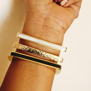 Puede incluir: Cuatro pulseras doradas apiladas en una muñeca. Las pulseras tienen diferentes diseños: una con una incrustación de esmalte blanco, otra con una superficie texturizada y otra con una incrustación de esmalte negro. Las uñas están pintadas con un diseño dorado y rosa.