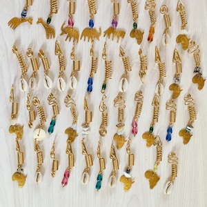 May include: An assortment of gold-coloured hair accessories, including spirals, charms, and beads. The charms feature animal shapes, seashells, and African continent outlines. They are arranged on a light wood-grain surface.