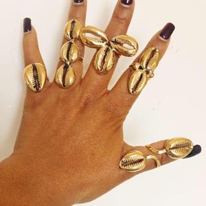 May include: A collection of gold-toned rings featuring cowrie shell designs worn on the fingers. The rings include single shell settings, a triple shell cluster, and multi-shell bands, all crafted with a polished metallic finish and textured details.