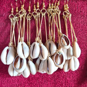 May include: A collection of gold-colored metal earrings featuring white cowrie shells. The earrings hang from gold-colored hooks and are displayed on a red fabric surface. The shells have a natural, organic shape.