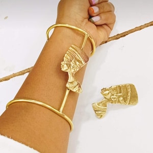 Op de afbeelding: Goudkleurige armband en hanger met een buste van Nefertiti. De armband is een eenvoudige band met een verticale staaf die de hanger vasthoudt. Er is ook een bijpassende hanger aanwezig. De sieraden worden op een lichte achtergrond getoond.