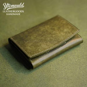 May include: Olive green leather wallet with a textured finish. The wallet has a flap closure and is stitched with a contrasting thread. The text "Ytcmould LEATHERGOODS HANDMADE" is visible in the upper left corner.