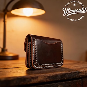 May include: A dark brown leather wallet with white stitching. The wallet has a curved flap and a pocket on the front. The text "LEATHERGOODS Ytcmould HANDMADE" is in the upper right corner. The wallet is on a wooden surface.