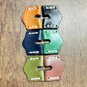 May include: Six leather watch strap keepers in tan, black, navy, green, olive, and brown. Each keeper is stitched with white thread and has a distinctive shape with a central opening. The keepers are arranged in a grid.